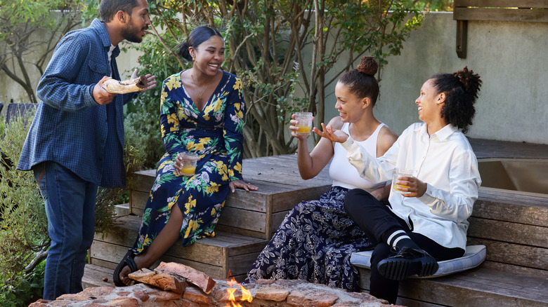 Smiling friends sitting and standing near a fire pit