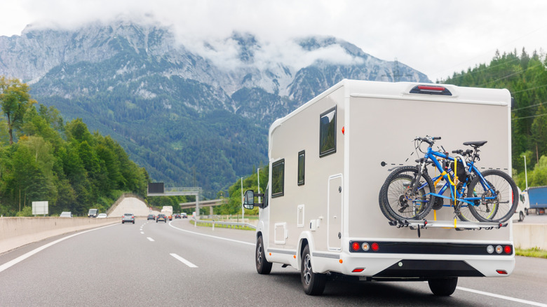 Class C RV with two bikes on the back driving down the road towards a mountain