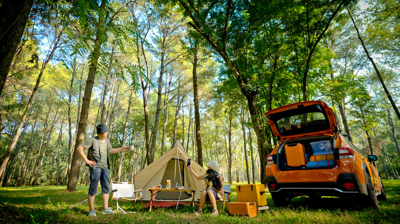 A man and woman camping in wooded area with a tent and orange SUV surrounded by tall trees.