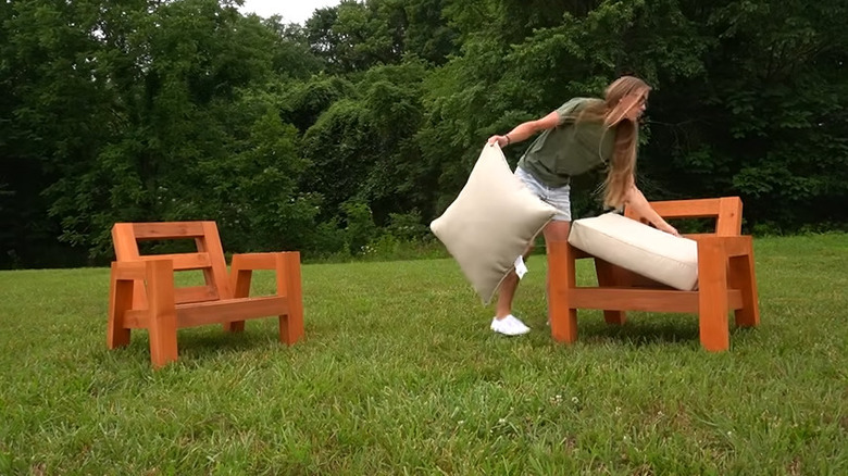 A woman placing large white pillows on a wooden chair in a grassy yard.