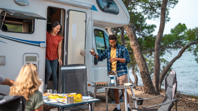 A group of friends crowd the outside of an RV, setting up a series of tables, chairs, and more