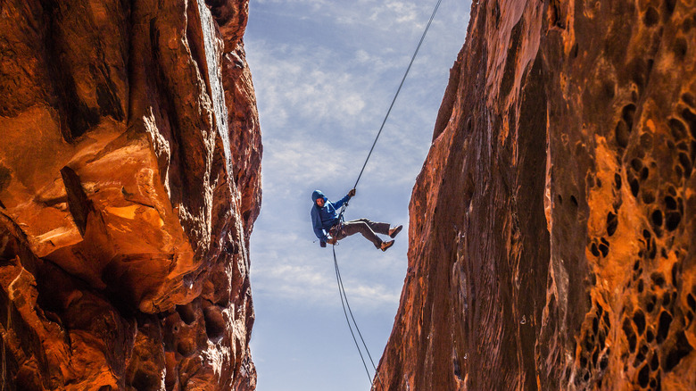 view from below of man in blue hooded jacket rappelling down red rock canyon