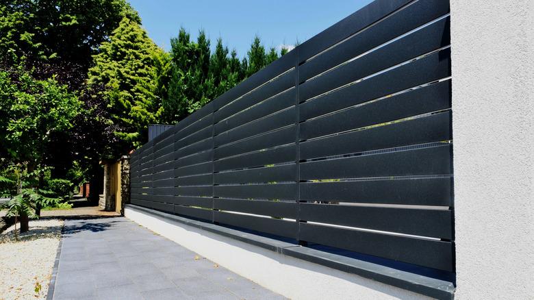 An extra-tall, dark gray aluminum privacy fence along a sidewalk