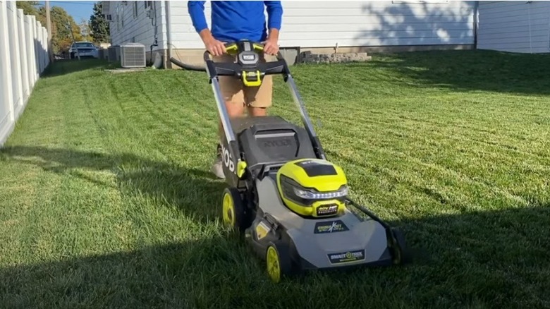 Man pushes Ryobi electric mower over green grass in small back yard.