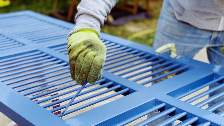 A person wearing gloves using blue paint and a small paintbrush on shutters