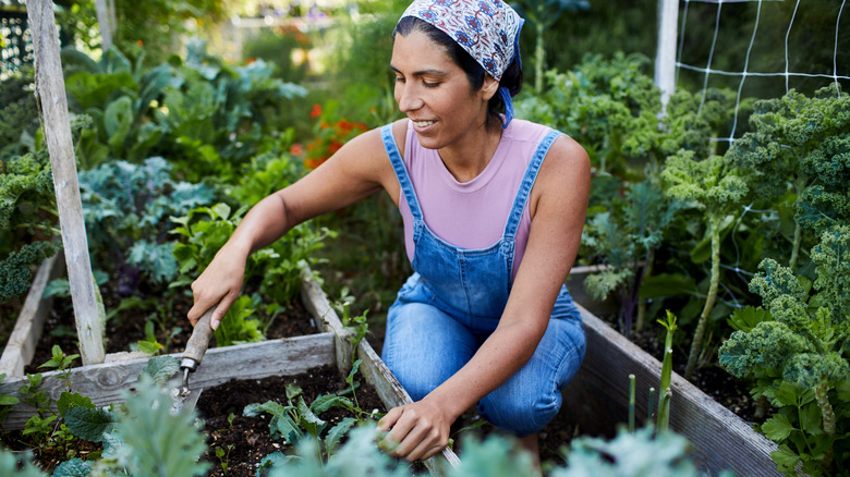 A woman working in garden in a pink shirt and denim overalls with a smile on her face.