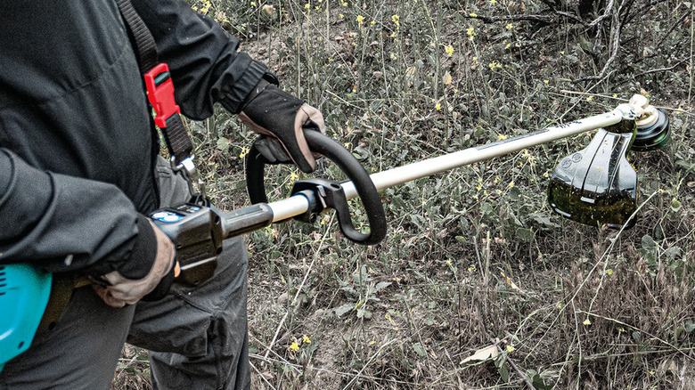 Figure in long sleeves and gloves holding a string trimmer in a weedy area