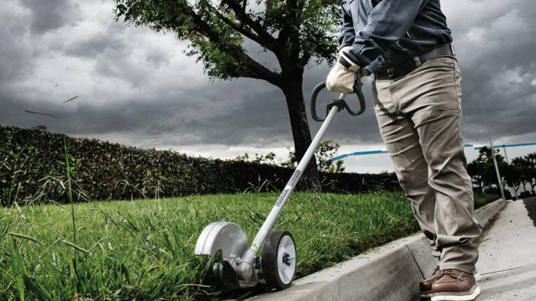 An angled view of a Makita edger along a curb, being held by a person in pants, long sleeves, and gloves