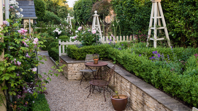 A rustic, French-inspired garden with stone wall, vintage bistro set, climbing flowers, hedges, and a gravel path.