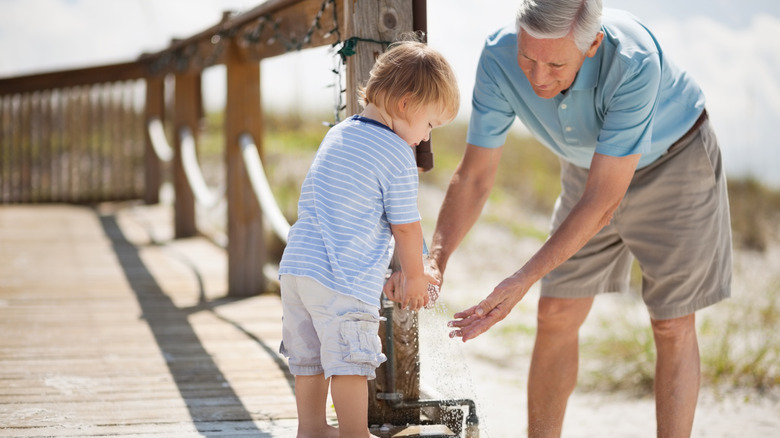 Grandfather helping young child wash hands at a faucet on a sandy shore