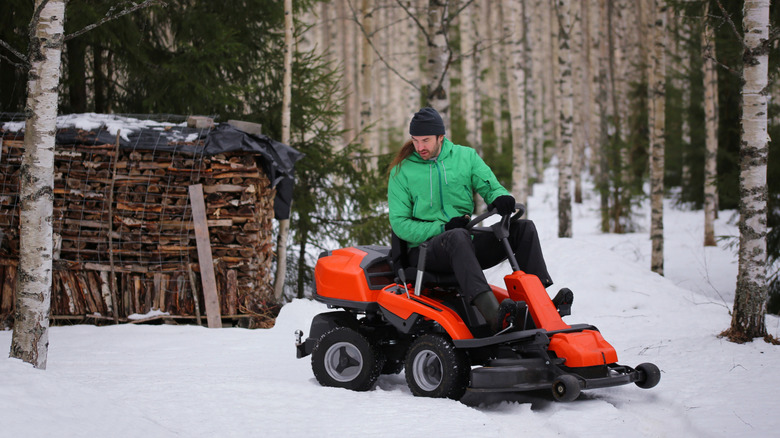 Man riding orange lawn mower in snow in forest and stacked firewood in background