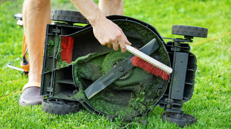 A person brushes out the caked-on grass under their lawn mower