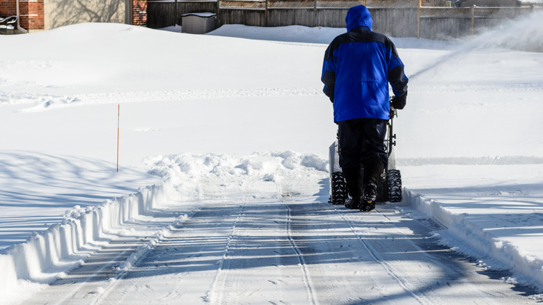 man uses snow blower to clear driveway, with driveway marker on one side