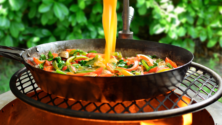 A cast iron skillet of vegetables and a stream of eggs yolks over an open flame