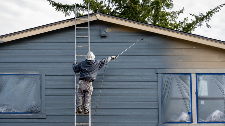 Worker on ladder painting house siding with a sprayer