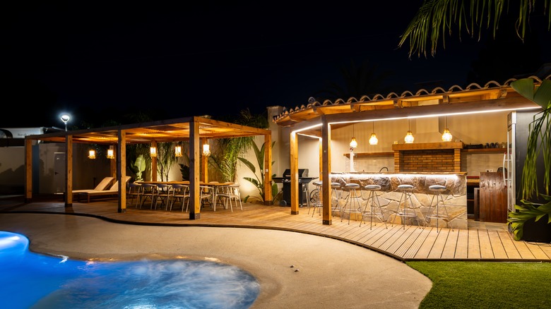 An outdoor pool, kitchen, and seating area cozily lit at night
