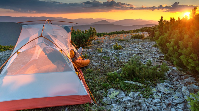 Person looking out of their tent at an orange sunset over distant mountains