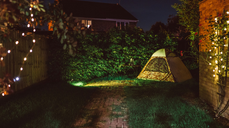 A tent in a backyard with a floodlight on it and string lights in trees in the foreground