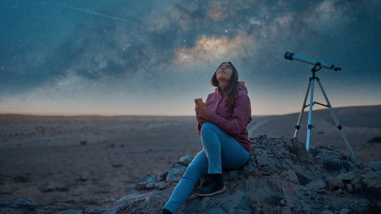 Woman sitting in the desert with a telescope, looking up at the night sky