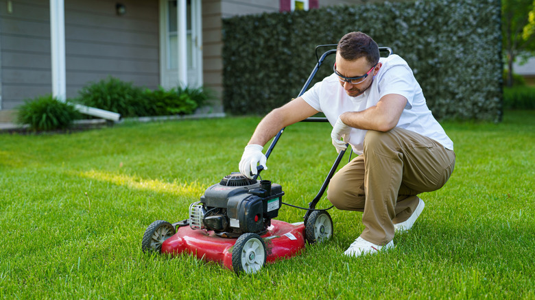 A homeowner kneels to inspect his lawnmower on the lawn