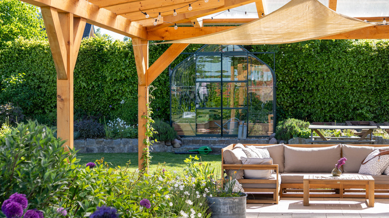 A covered patio in a lush backyard with a greenhouse in the background.