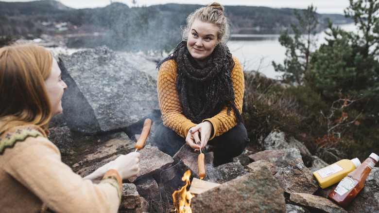 Two women cooking hot dogs over a rocky campfire while smiling with a lake in the background.