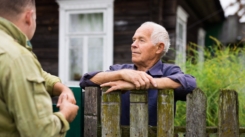 Two neighbors talking over their shared wooden fence.