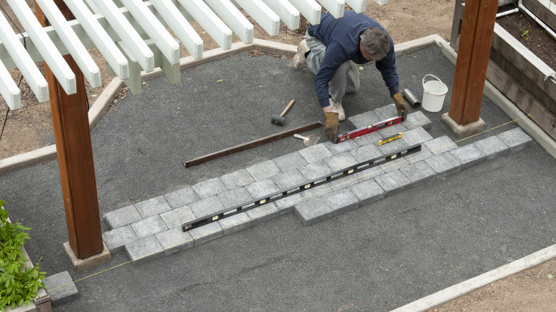 A man checking the level on a partial patio design with pavers on small stones under a pergola
